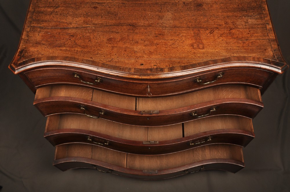 An 18th-century mahogany serpentine chest of drawers featuring four graduated drawers, carved canted corners, and brass swan-neck handles. View X