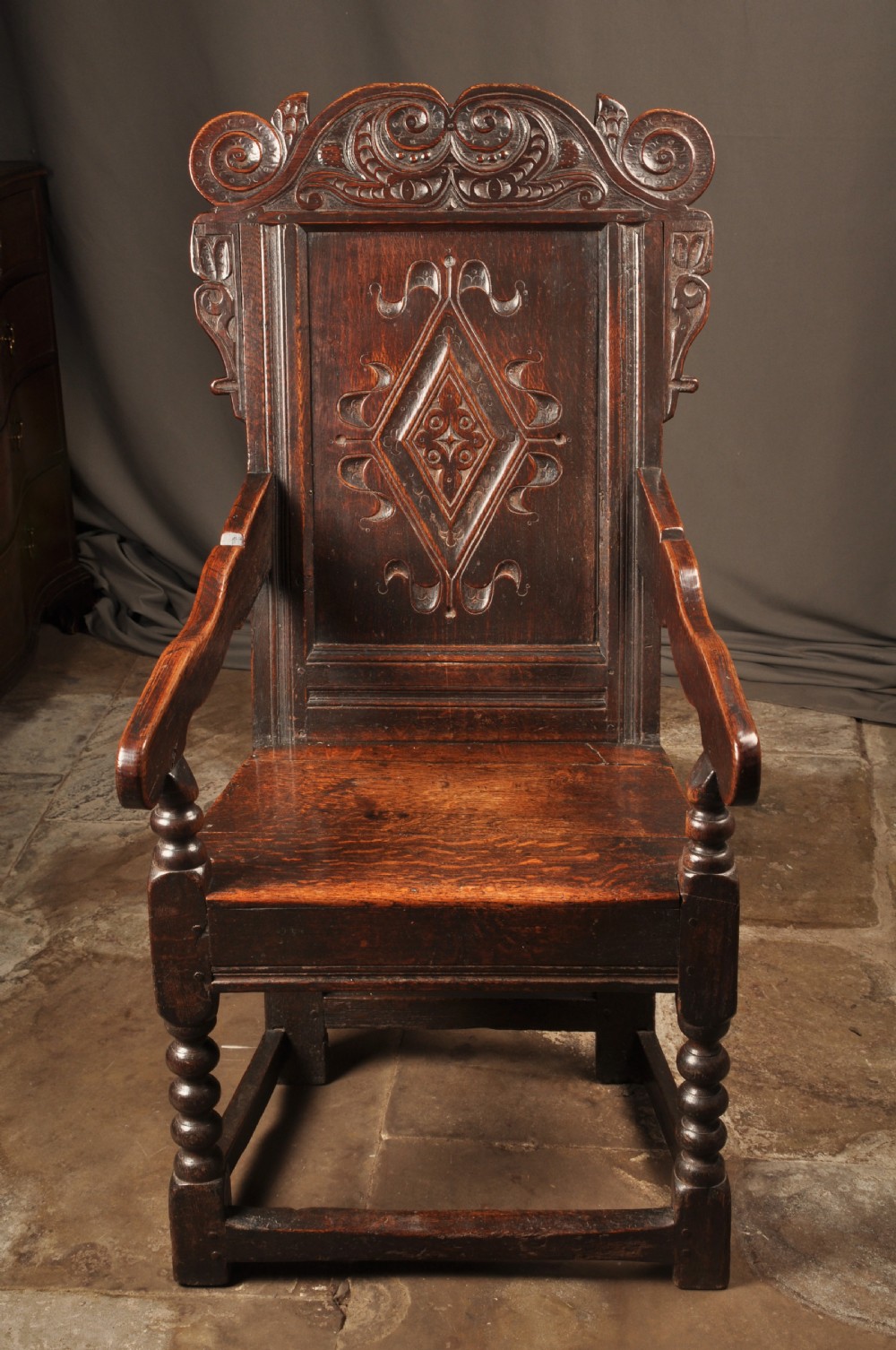 A 17th-century dark oak wainscot armchair featuring a carved scroll cresting, lozenge-carved back panel, shaped armrests, and turned front legs.