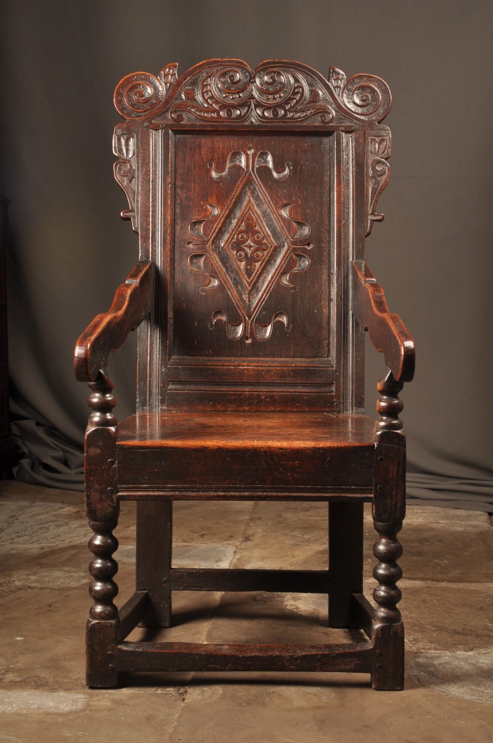 A 17th-century dark oak wainscot armchair featuring a carved scroll cresting, lozenge-carved back panel, shaped armrests, and turned front legs. View II