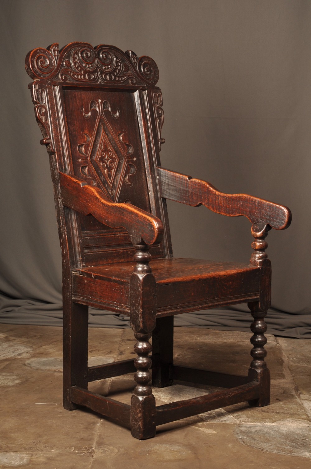 A 17th-century dark oak wainscot armchair featuring a carved scroll cresting, lozenge-carved back panel, shaped armrests, and turned front legs. View VI