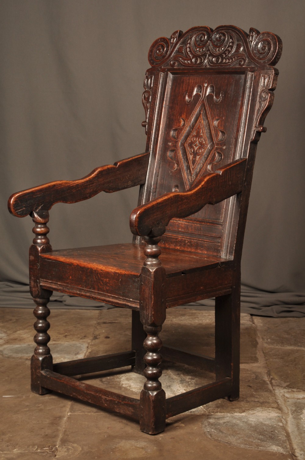 A 17th-century dark oak wainscot armchair featuring a carved scroll cresting, lozenge-carved back panel, shaped armrests, and turned front legs. View VII