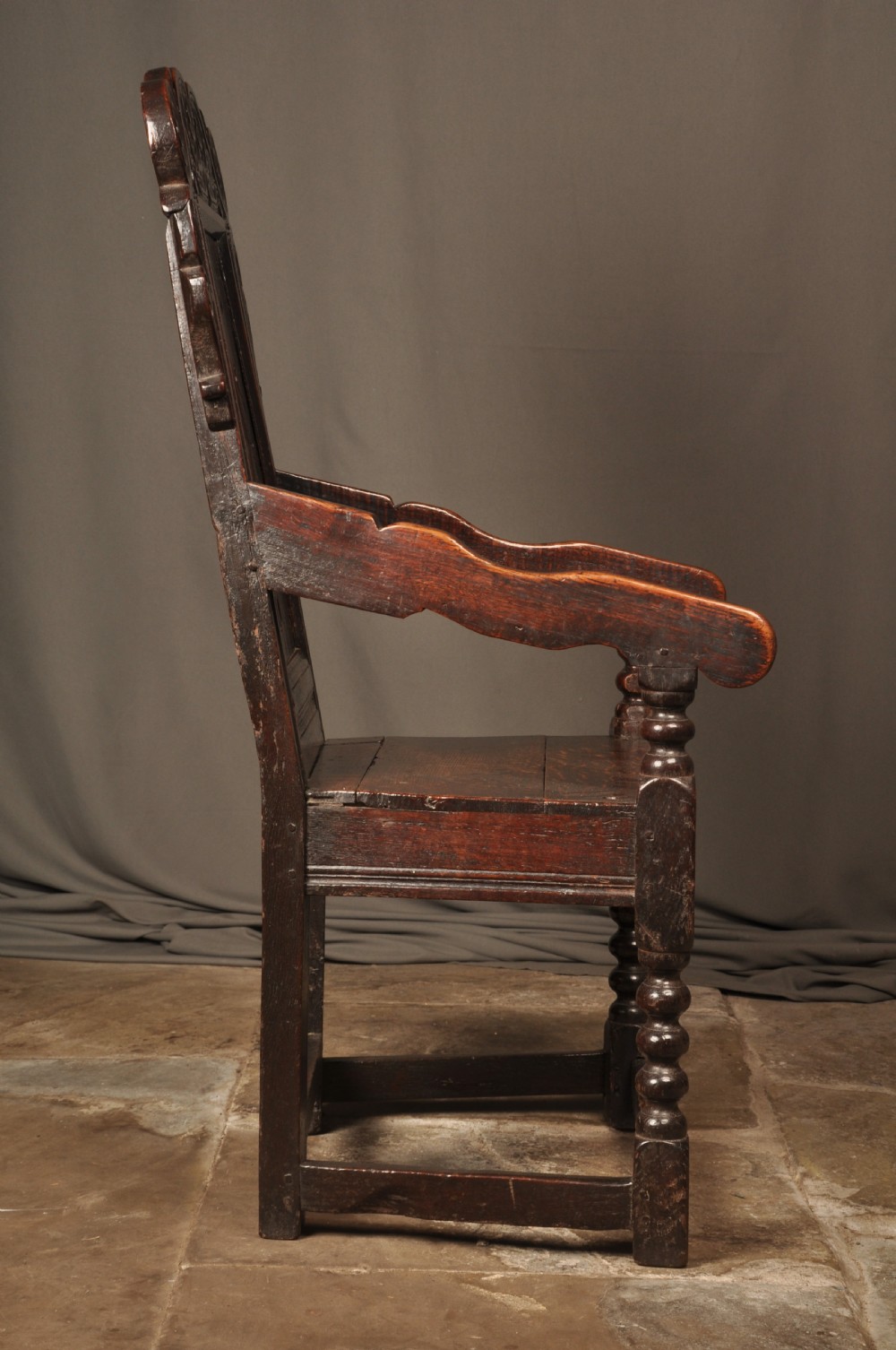 A 17th-century dark oak wainscot armchair featuring a carved scroll cresting, lozenge-carved back panel, shaped armrests, and turned front legs. View VIII