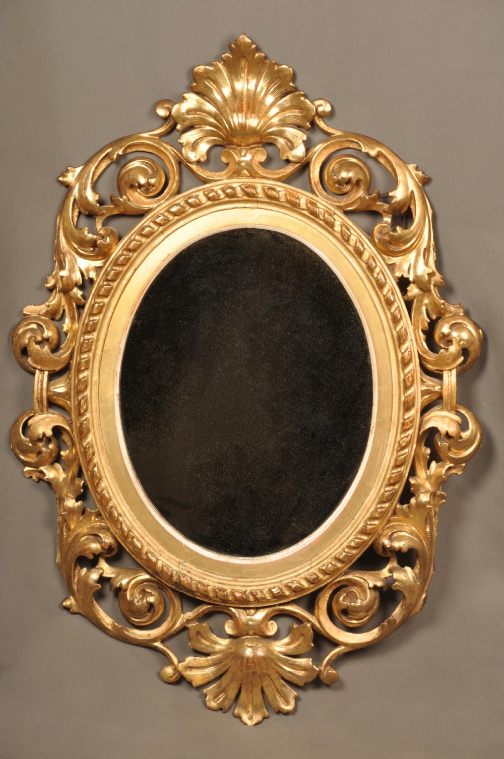 An antique oval giltwood wall mirror featuring an intricately carved openwork frame decorated with scrolling acanthus leaves, C-scrolls, rope moulding, and shell cresting.
