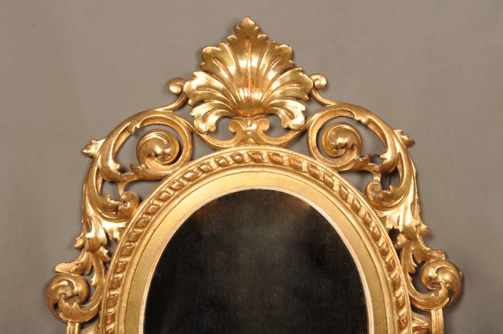 An antique oval giltwood wall mirror featuring an intricately carved openwork frame decorated with scrolling acanthus leaves, C-scrolls, rope moulding, and shell cresting. View II