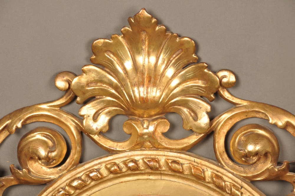 An antique oval giltwood wall mirror featuring an intricately carved openwork frame decorated with scrolling acanthus leaves, C-scrolls, rope moulding, and shell cresting. View III