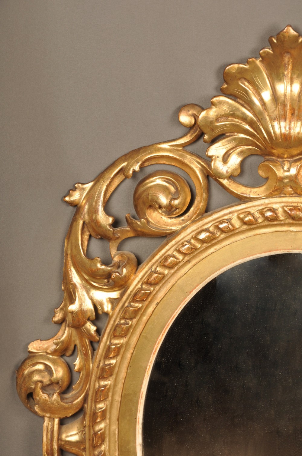 An antique oval giltwood wall mirror featuring an intricately carved openwork frame decorated with scrolling acanthus leaves, C-scrolls, rope moulding, and shell cresting. View IV