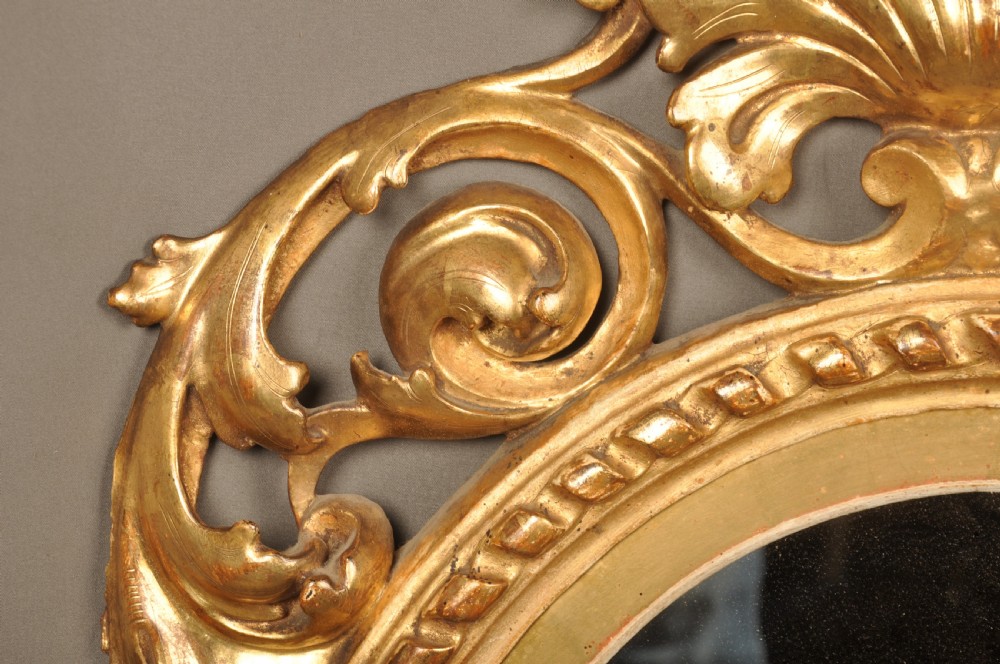 An antique oval giltwood wall mirror featuring an intricately carved openwork frame decorated with scrolling acanthus leaves, C-scrolls, rope moulding, and shell cresting. View V