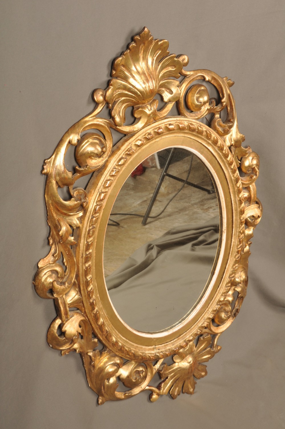 An antique oval giltwood wall mirror featuring an intricately carved openwork frame decorated with scrolling acanthus leaves, C-scrolls, rope moulding, and shell cresting. View VI