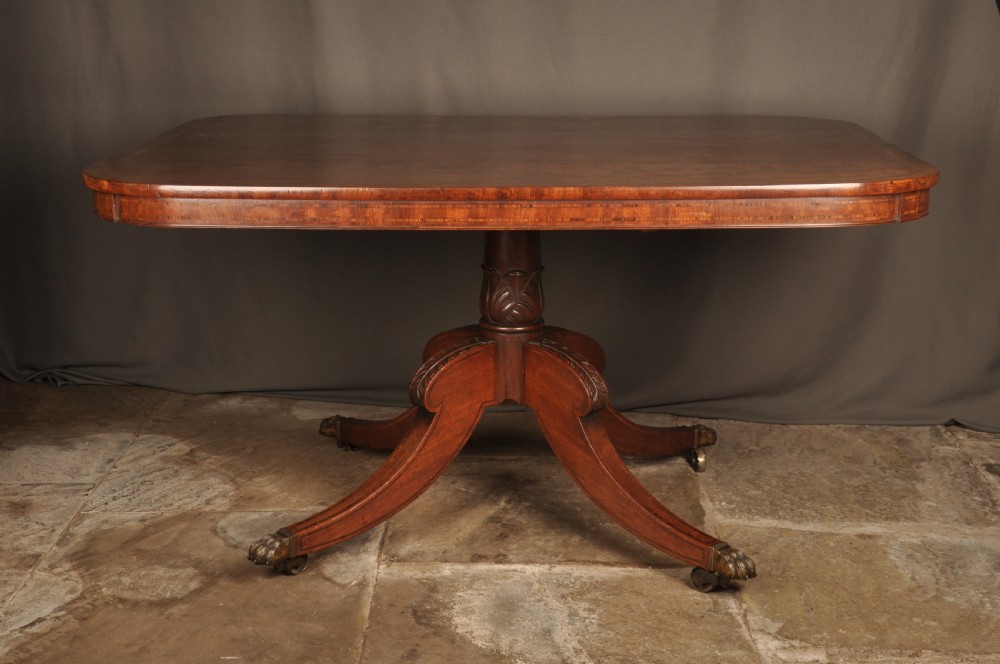A Regency mahogany breakfast table featuring a rounded rectangular top, a turned central pedestal with acanthus carving, and four splayed legs with brass paw feet.