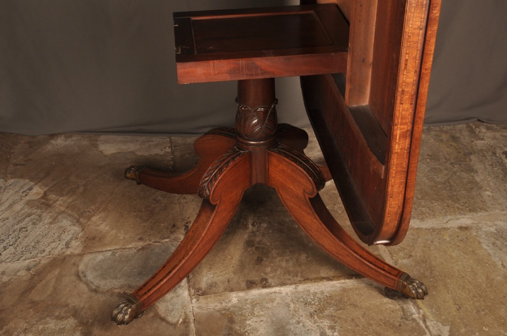 A Regency mahogany breakfast table featuring a rounded rectangular top, a turned central pedestal with acanthus carving, and four splayed legs with brass paw feet. View IX