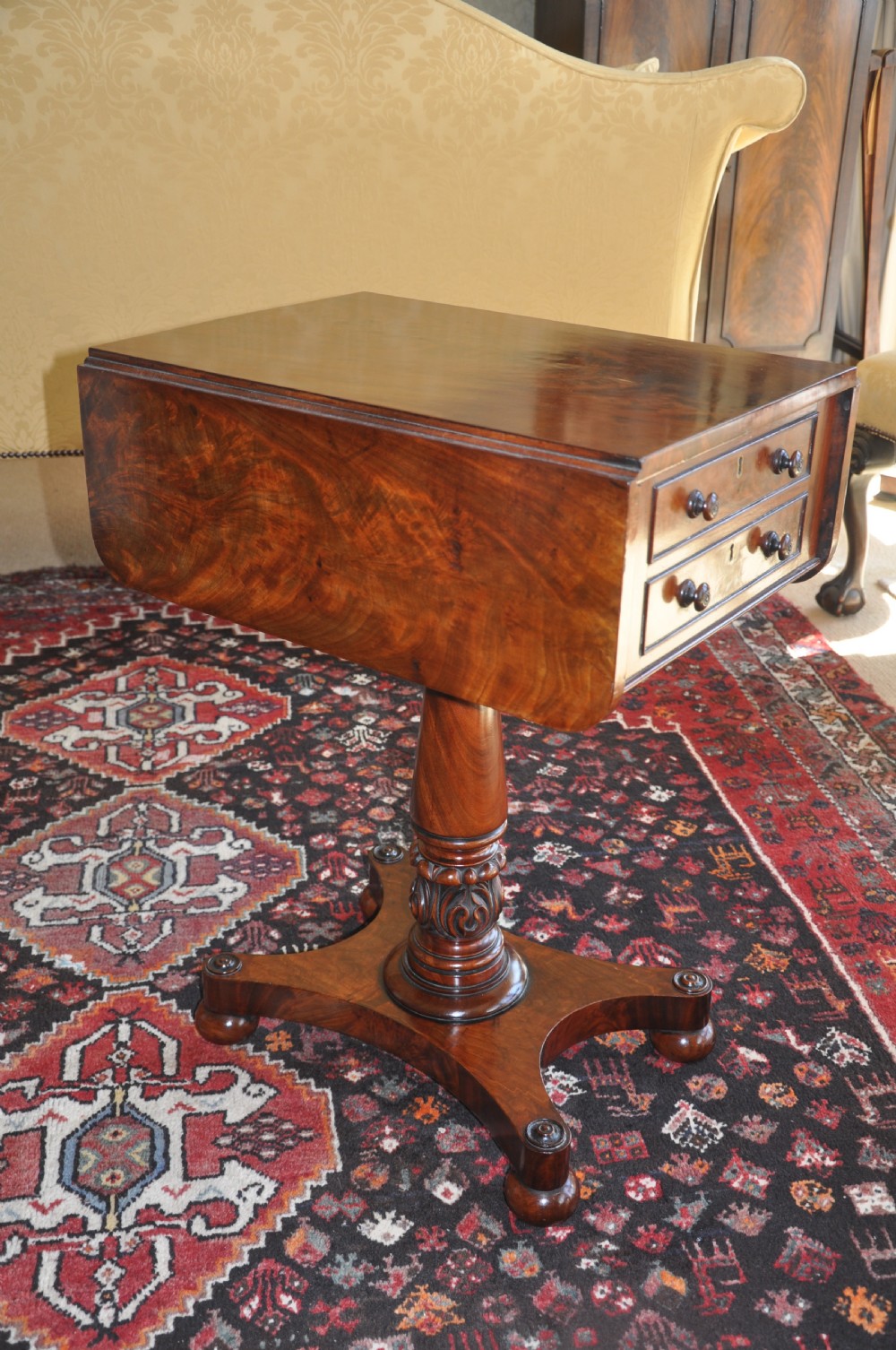 A Regency flame mahogany Pembroke table featuring two drop leaves, two small drawers with turned wooden handles, a carved turned pedestal support, and a quadruped platform base. View II