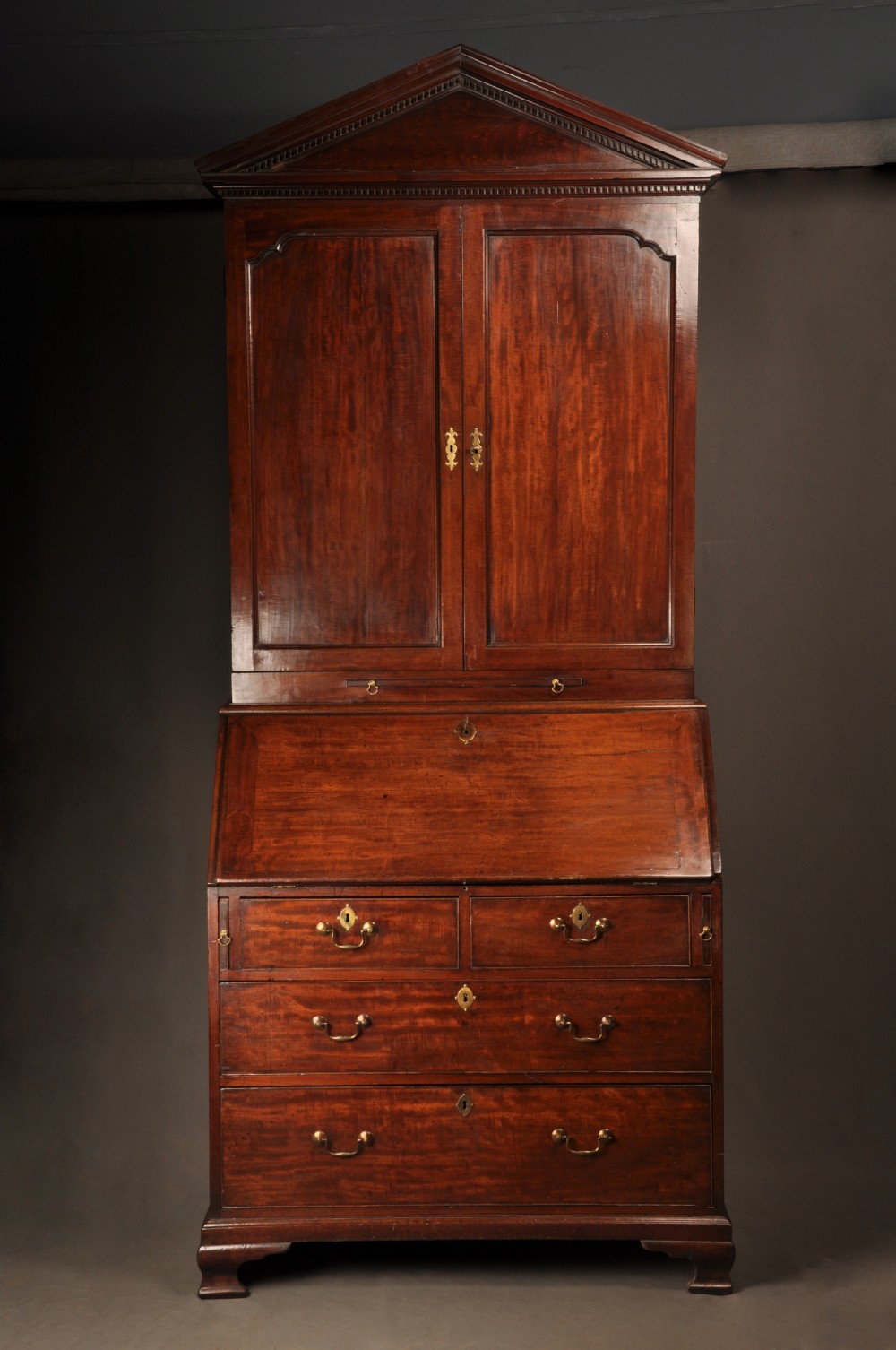An 18th-century mahogany bureau cabinet featuring an architectural pediment top, two panelled upper doors, a slant-front writing desk, and lower drawers on bracket feet.