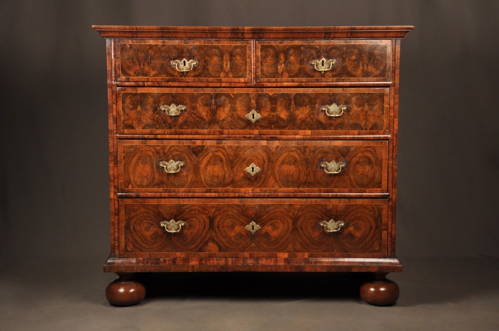 A seventeenth-century olive oyster veneered chest of drawers featuring two short drawers above three long drawers, brass drop handles, and turned bun feet.