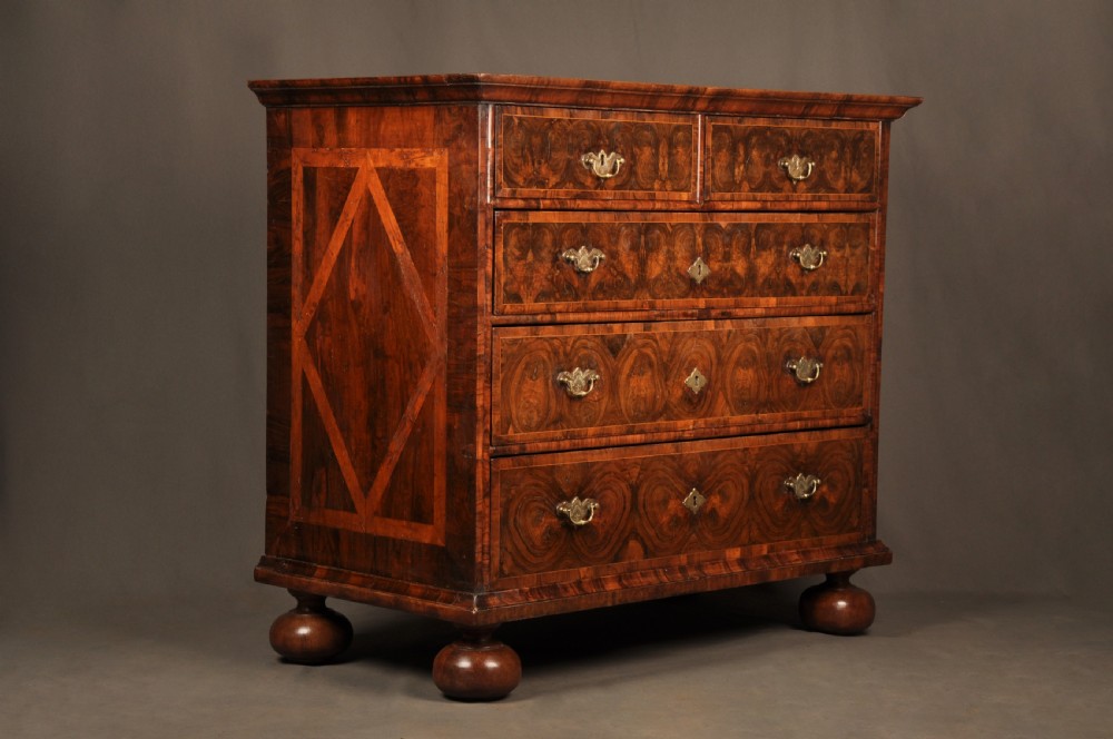 A seventeenth-century olive oyster veneered chest of drawers featuring two short drawers above three long drawers, brass drop handles, and turned bun feet. View II