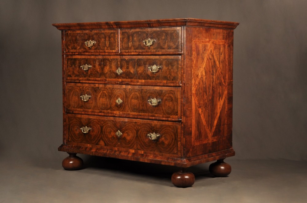 A seventeenth-century olive oyster veneered chest of drawers featuring two short drawers above three long drawers, brass drop handles, and turned bun feet. View III