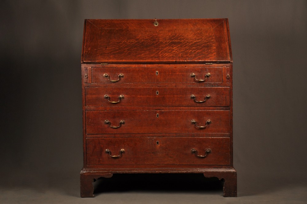 A mid-18th-century warm brown oak slant-front writing bureau featuring four graduated drawers with brass swan-neck handles and bracket feet.