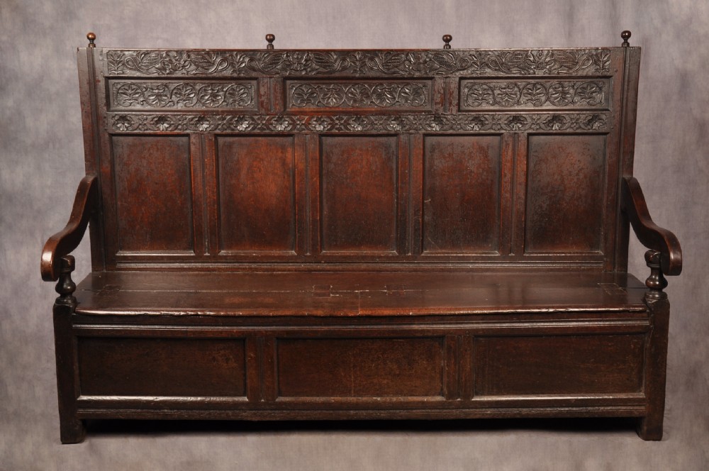 A dark oak 17th-century box settle featuring a high panelled back with carved floral friezes, turned finials, shaped armrests, and a lifted seat exposing a storage base.
