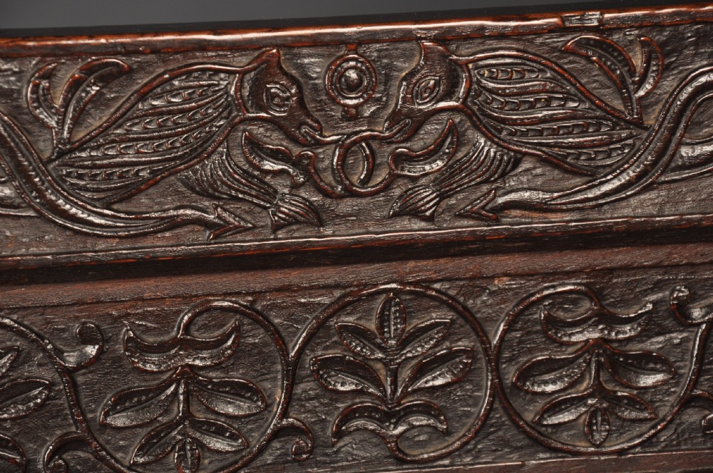 A dark oak 17th-century box settle featuring a high panelled back with carved floral friezes, turned finials, shaped armrests, and a lifted seat exposing a storage base. View II