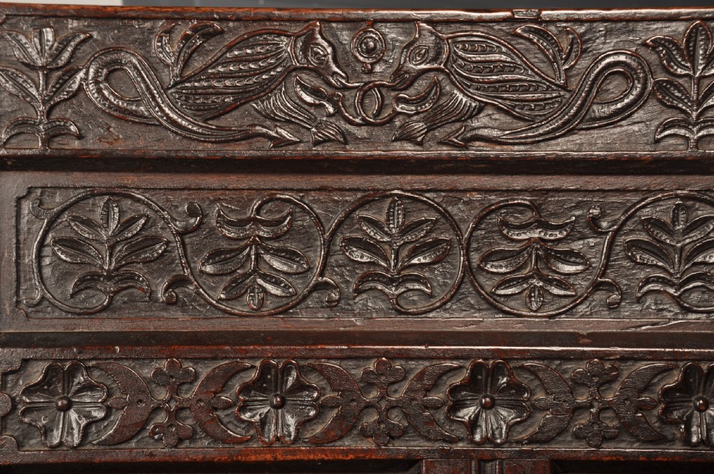 A dark oak 17th-century box settle featuring a high panelled back with carved floral friezes, turned finials, shaped armrests, and a lifted seat exposing a storage base. View III
