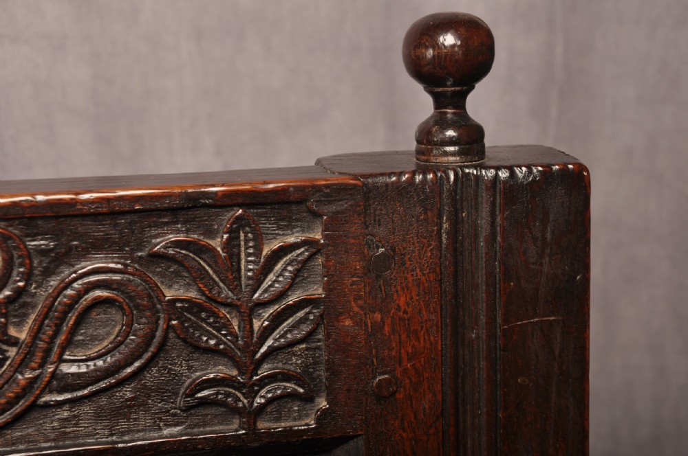 A dark oak 17th-century box settle featuring a high panelled back with carved floral friezes, turned finials, shaped armrests, and a lifted seat exposing a storage base. View V