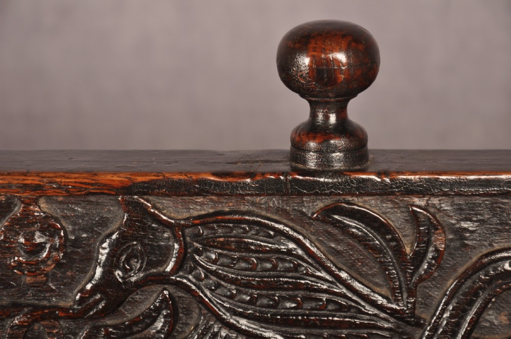 A dark oak 17th-century box settle featuring a high panelled back with carved floral friezes, turned finials, shaped armrests, and a lifted seat exposing a storage base. View VI