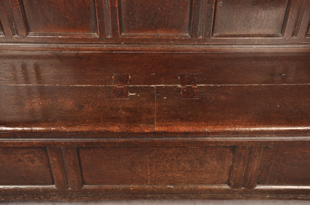 A dark oak 17th-century box settle featuring a high panelled back with carved floral friezes, turned finials, shaped armrests, and a lifted seat exposing a storage base. View VII