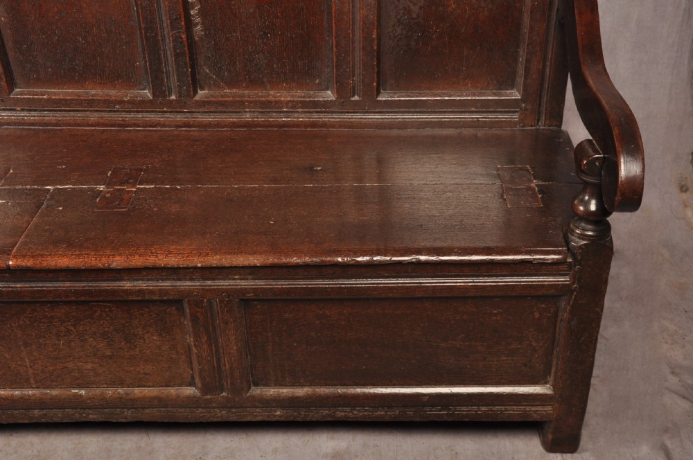 A dark oak 17th-century box settle featuring a high panelled back with carved floral friezes, turned finials, shaped armrests, and a lifted seat exposing a storage base. View VIII