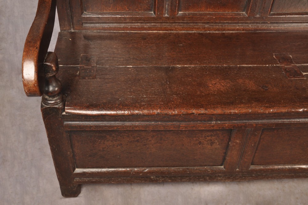 A dark oak 17th-century box settle featuring a high panelled back with carved floral friezes, turned finials, shaped armrests, and a lifted seat exposing a storage base. View IX