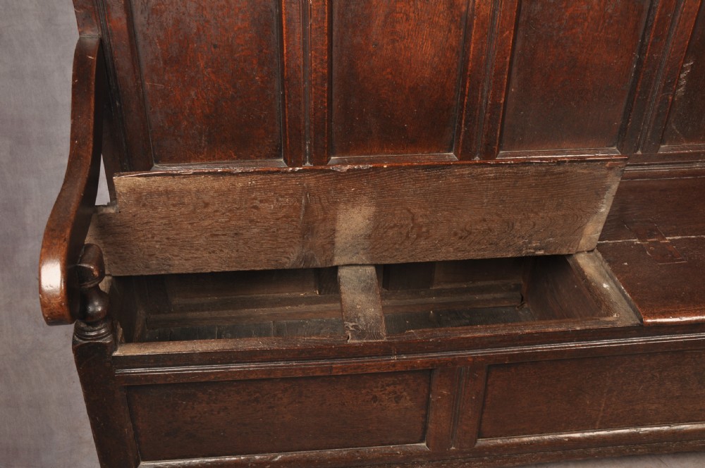 A dark oak 17th-century box settle featuring a high panelled back with carved floral friezes, turned finials, shaped armrests, and a lifted seat exposing a storage base. View X