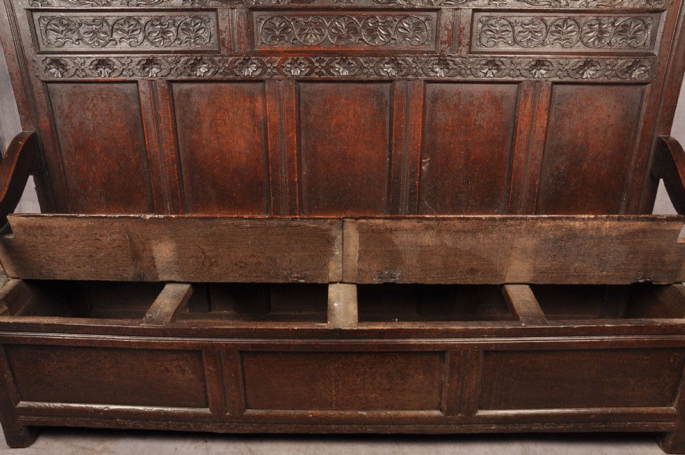 A dark oak 17th-century box settle featuring a high panelled back with carved floral friezes, turned finials, shaped armrests, and a lifted seat exposing a storage base. View XI