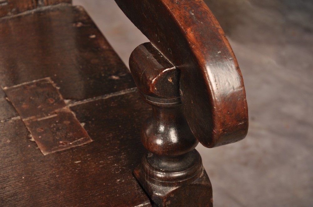 A dark oak 17th-century box settle featuring a high panelled back with carved floral friezes, turned finials, shaped armrests, and a lifted seat exposing a storage base. View XII