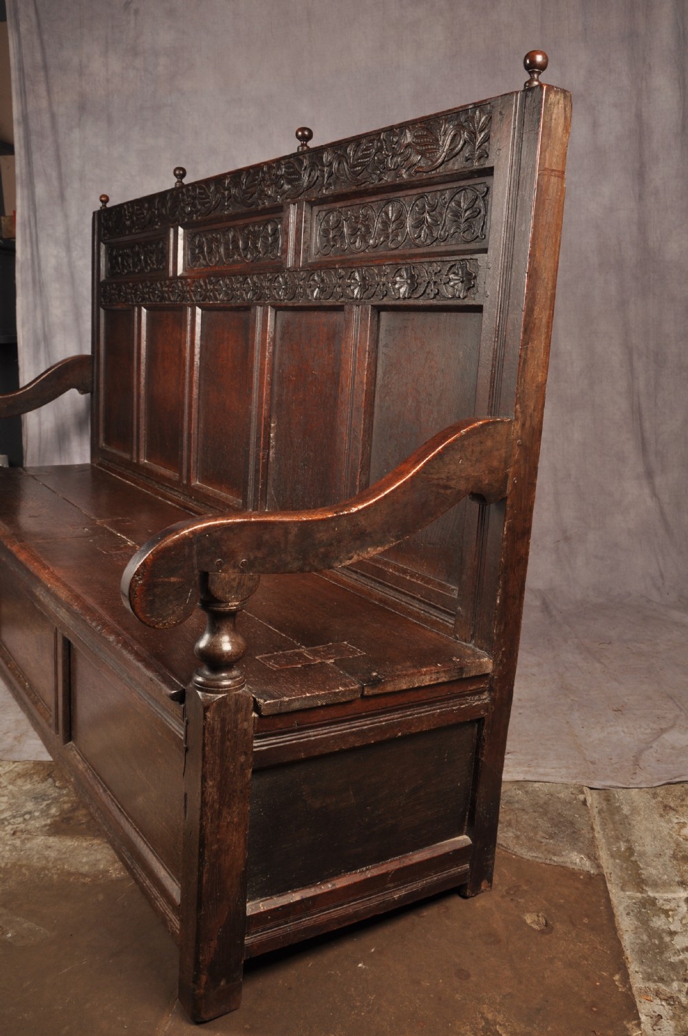A dark oak 17th-century box settle featuring a high panelled back with carved floral friezes, turned finials, shaped armrests, and a lifted seat exposing a storage base. View XIII