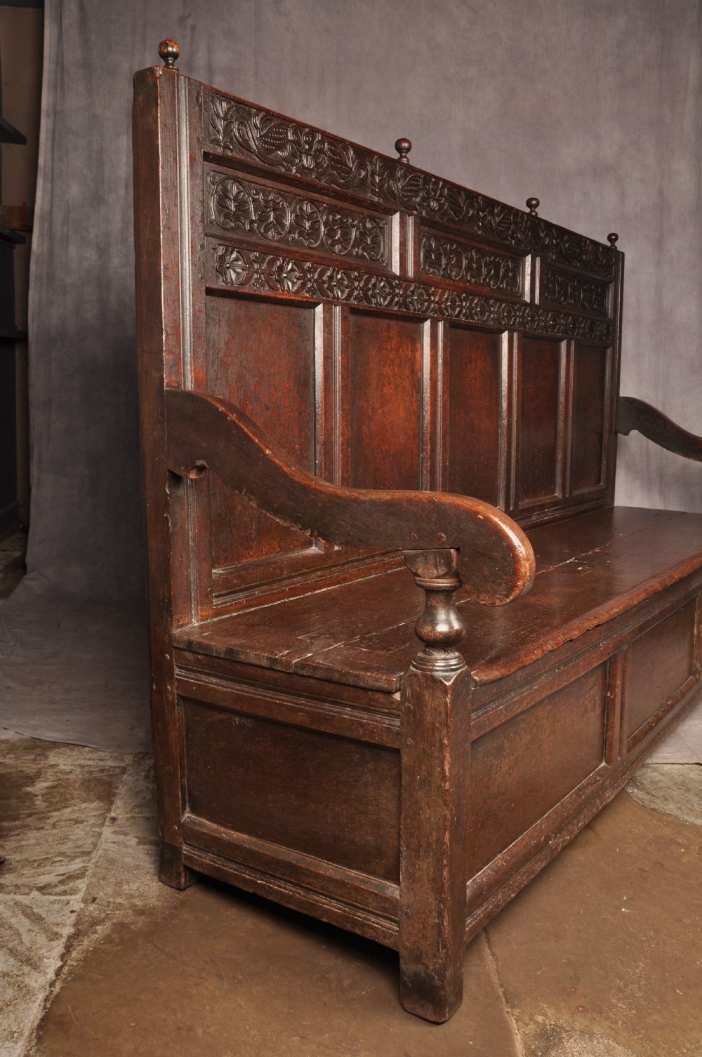 A dark oak 17th-century box settle featuring a high panelled back with carved floral friezes, turned finials, shaped armrests, and a lifted seat exposing a storage base. View XIV