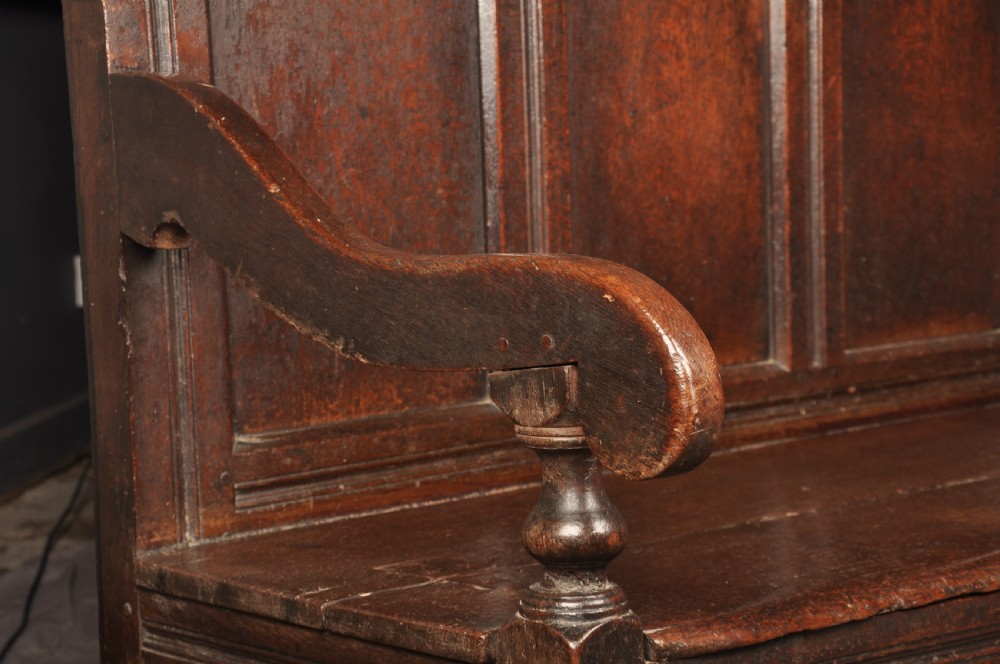 A dark oak 17th-century box settle featuring a high panelled back with carved floral friezes, turned finials, shaped armrests, and a lifted seat exposing a storage base. View XV