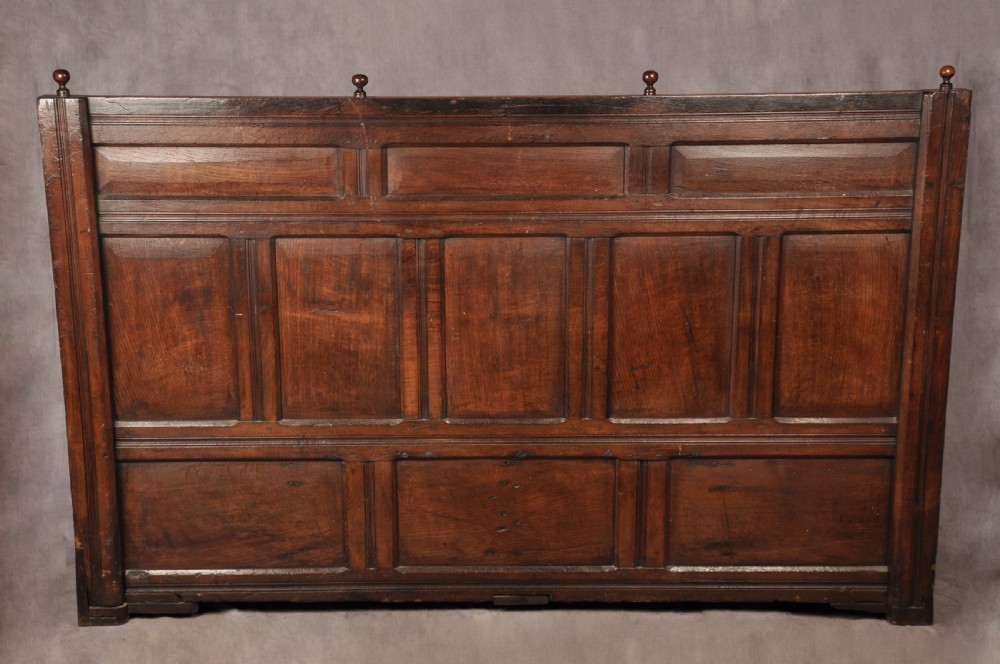 A dark oak 17th-century box settle featuring a high panelled back with carved floral friezes, turned finials, shaped armrests, and a lifted seat exposing a storage base. View XVI