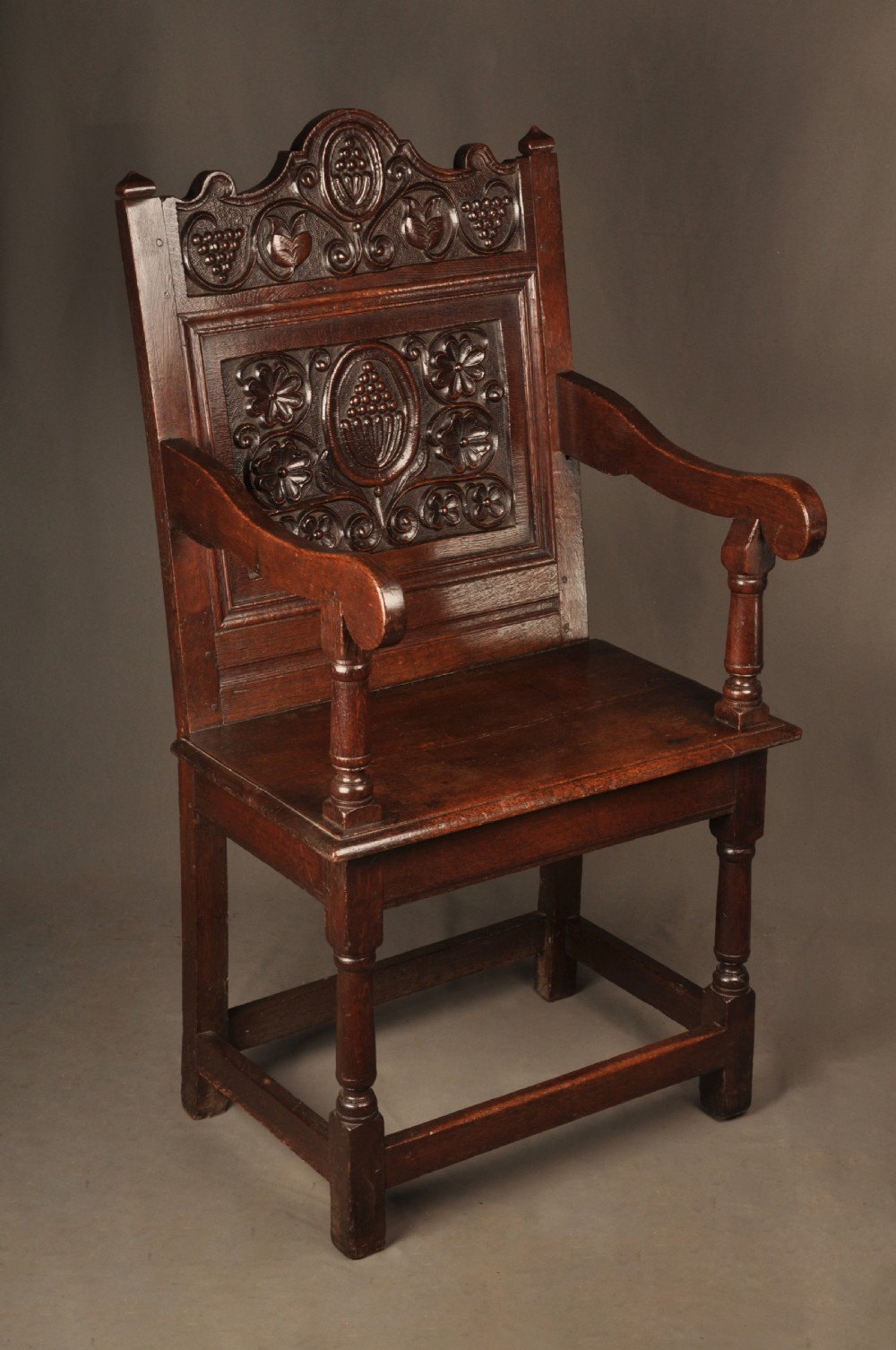 A seventeenth-century dark oak wainscot armchair featuring a carved back panel with floral motifs, turned supports, and shaped armrests.
