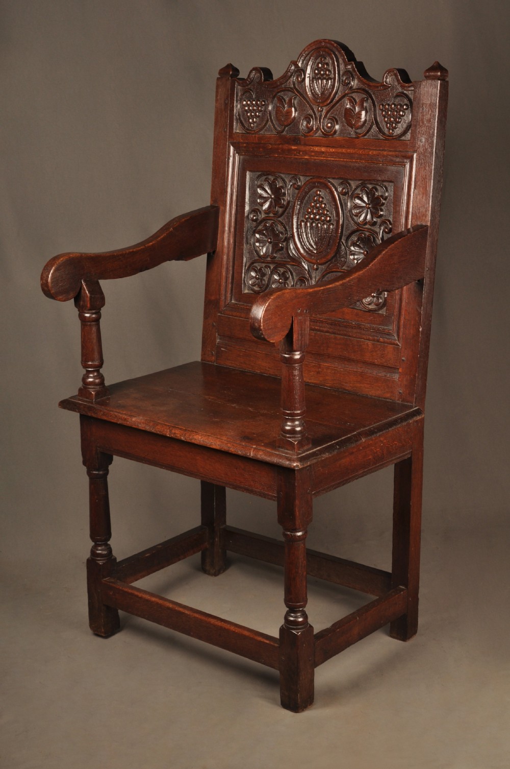 A seventeenth-century dark oak wainscot armchair featuring a carved back panel with floral motifs, turned supports, and shaped armrests. View II