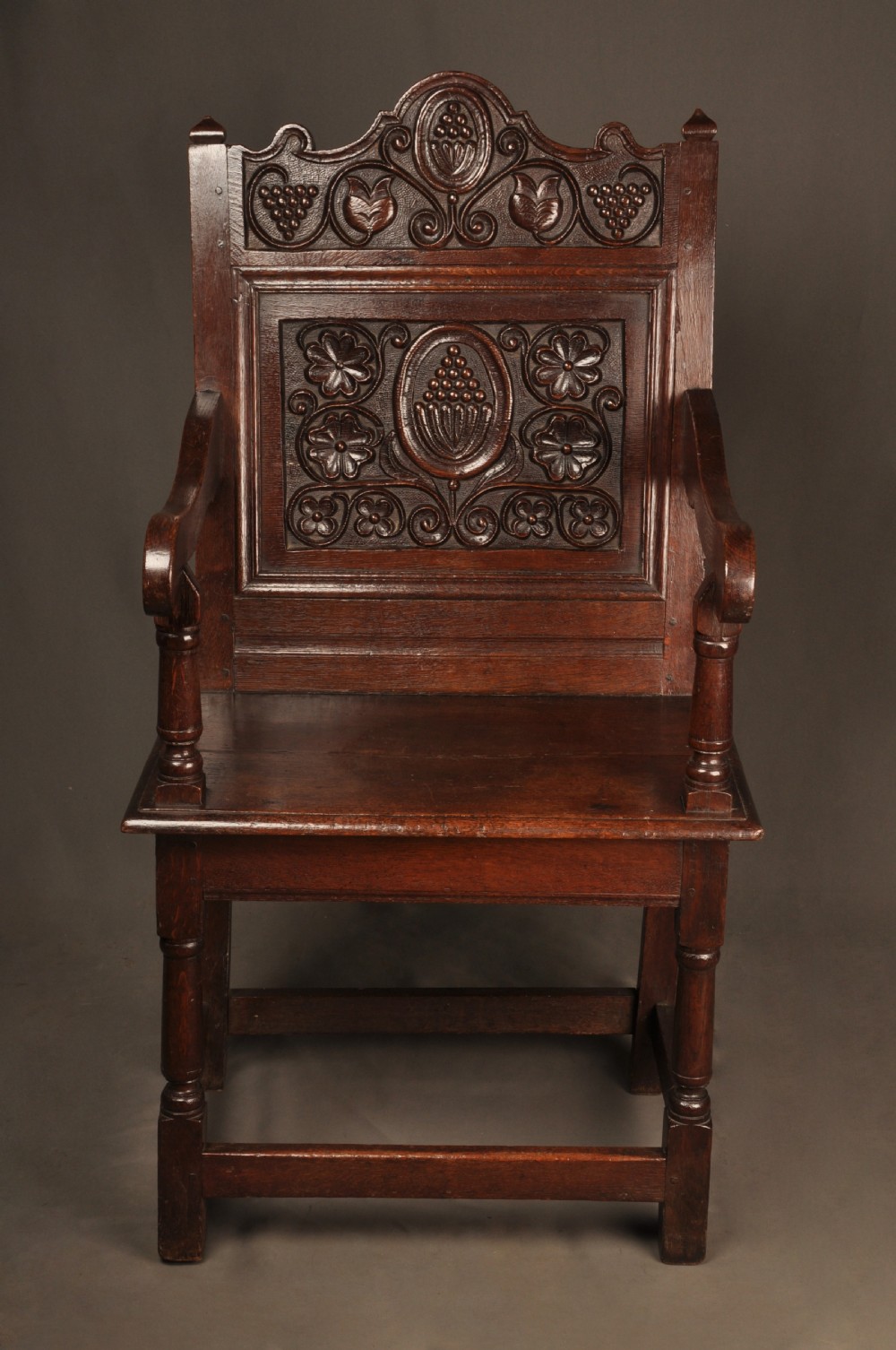A seventeenth-century dark oak wainscot armchair featuring a carved back panel with floral motifs, turned supports, and shaped armrests. View III