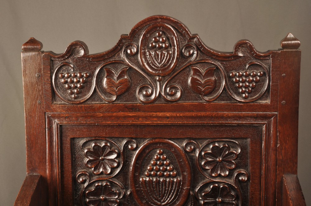 A seventeenth-century dark oak wainscot armchair featuring a carved back panel with floral motifs, turned supports, and shaped armrests. View V
