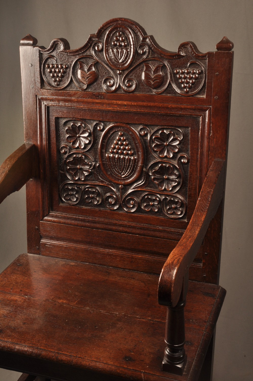 A seventeenth-century dark oak wainscot armchair featuring a carved back panel with floral motifs, turned supports, and shaped armrests. View X