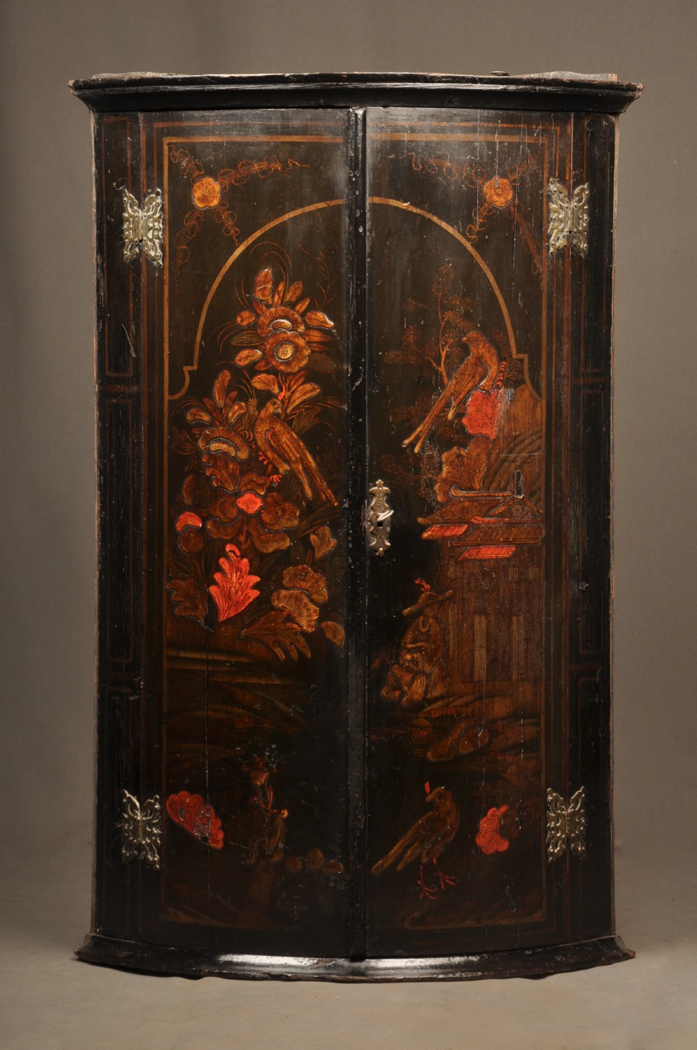 An early 18th-century black japanned hanging corner cupboard featuring hand-painted chinoiserie decoration depicting birds, flora, and architectural structures with brass butterfly hinges.