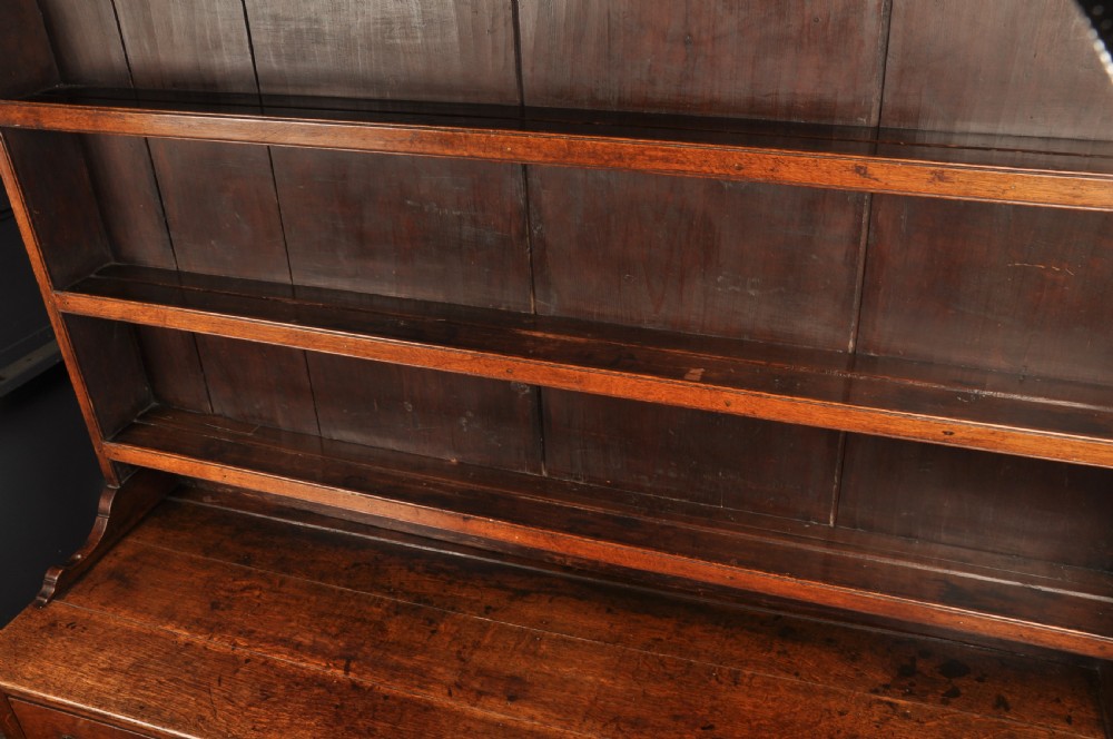 An eighteenth-century warm brown oak dresser featuring an upper three-tier open plate rack above a lower cabinet with drawers and panelled cupboard doors. View XIV