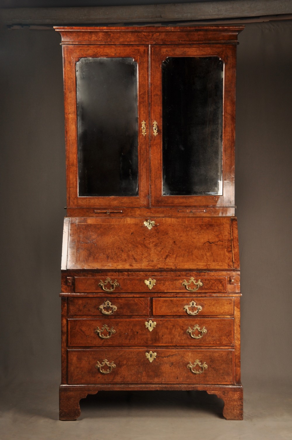 A George I burr walnut bureau bookcase with double mirrored upper doors over a slant-front slope and chest of drawers with brass handles.