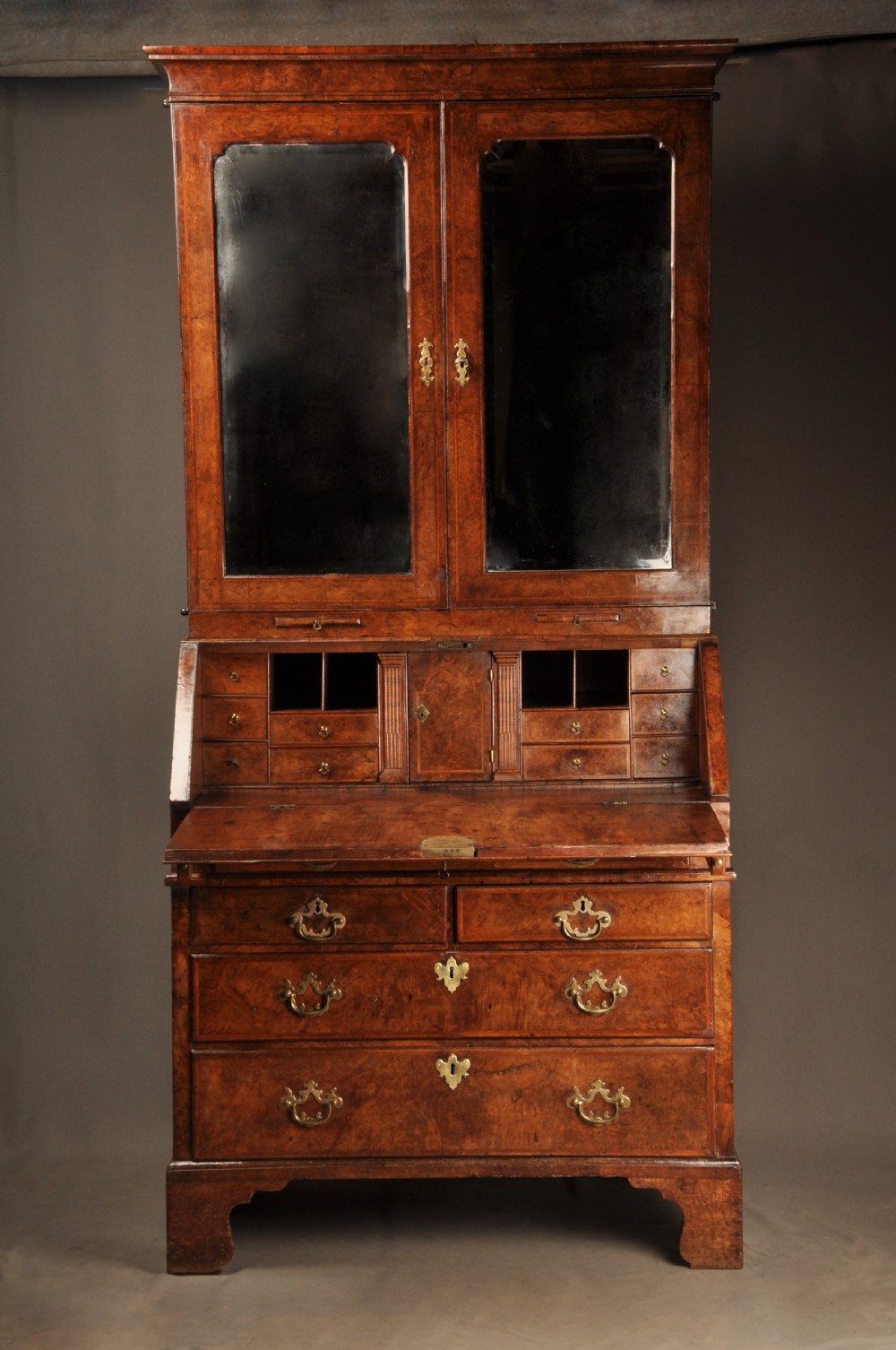 A George I burr walnut bureau bookcase with double mirrored upper doors over a slant-front slope and chest of drawers with brass handles. View XIV
