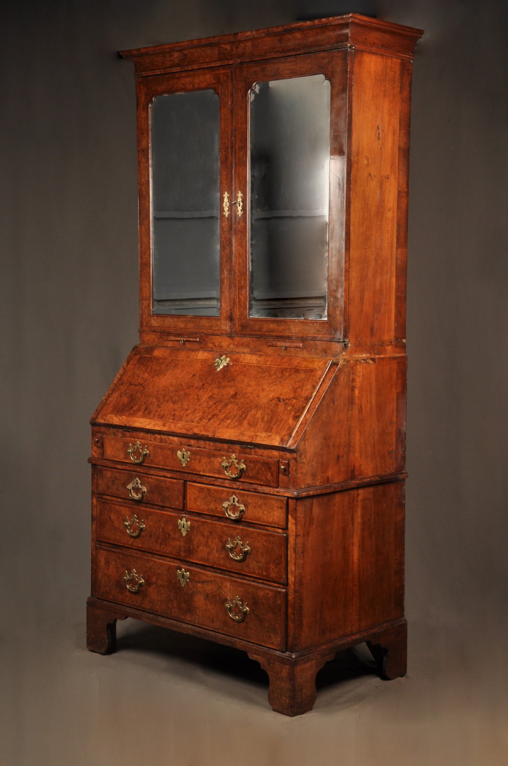A George I burr walnut bureau bookcase with double mirrored upper doors over a slant-front slope and chest of drawers with brass handles. View XV