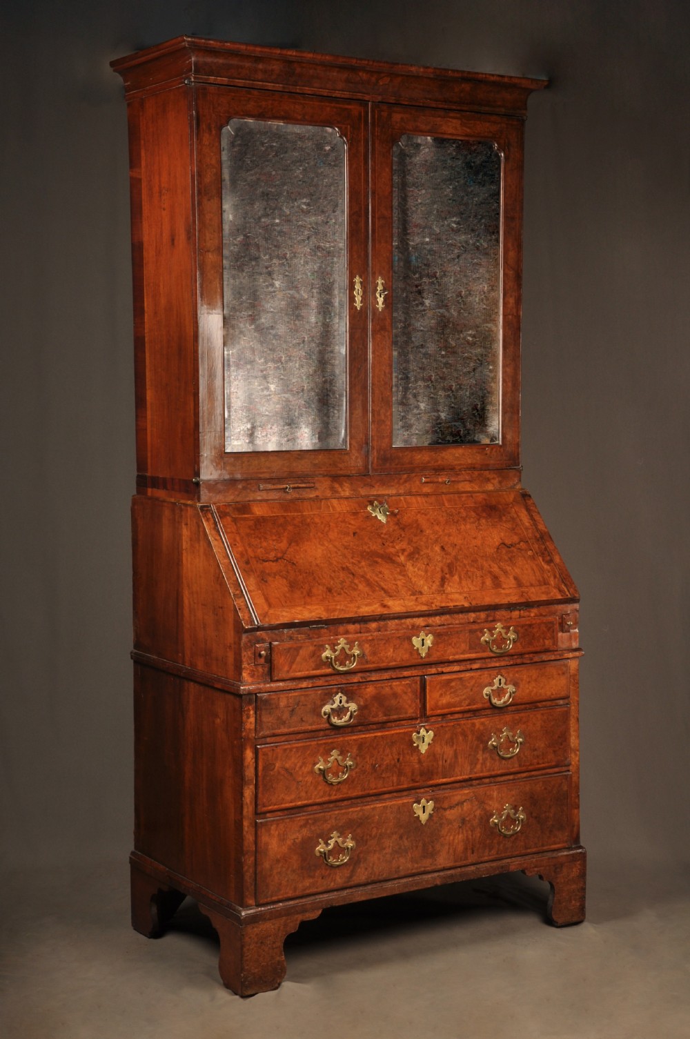 A George I burr walnut bureau bookcase with double mirrored upper doors over a slant-front slope and chest of drawers with brass handles. View XVI