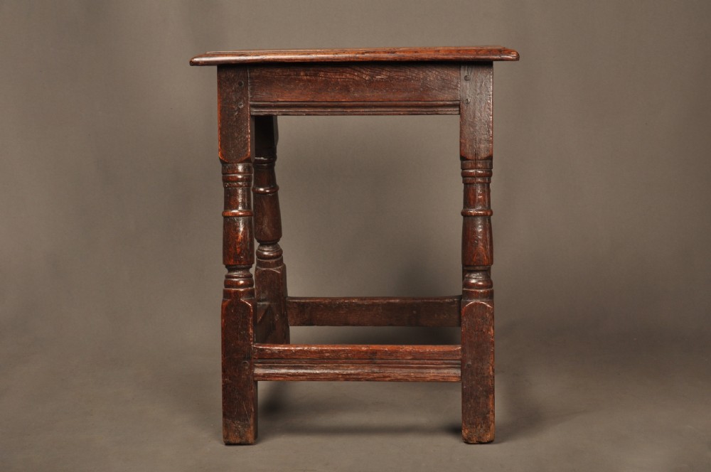 An 18th-century oak joint stool featuring baluster turned legs, plain stretchers, a rectangular moulded top, and dark brown patinated wood.
