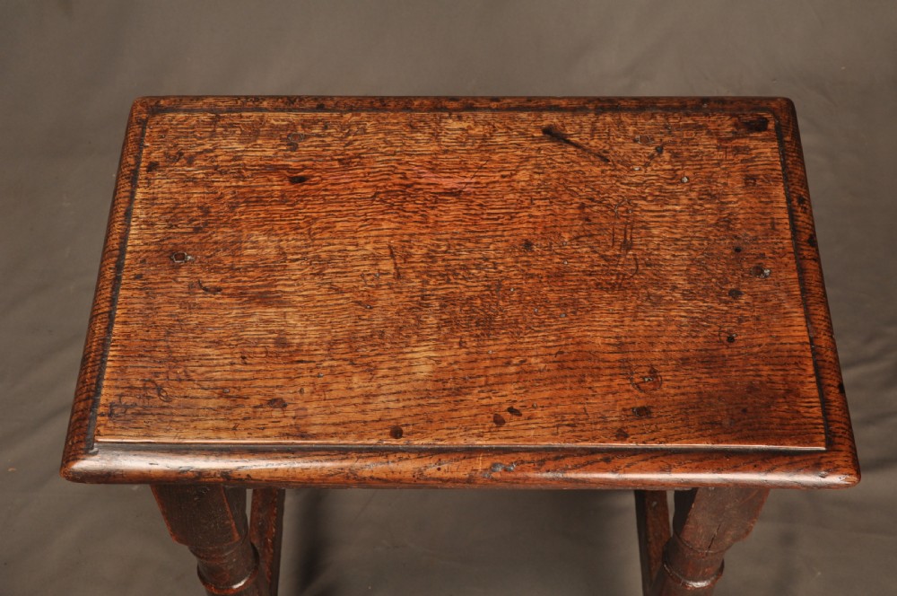 An 18th-century oak joint stool featuring baluster turned legs, plain stretchers, a rectangular moulded top, and dark brown patinated wood. View X
