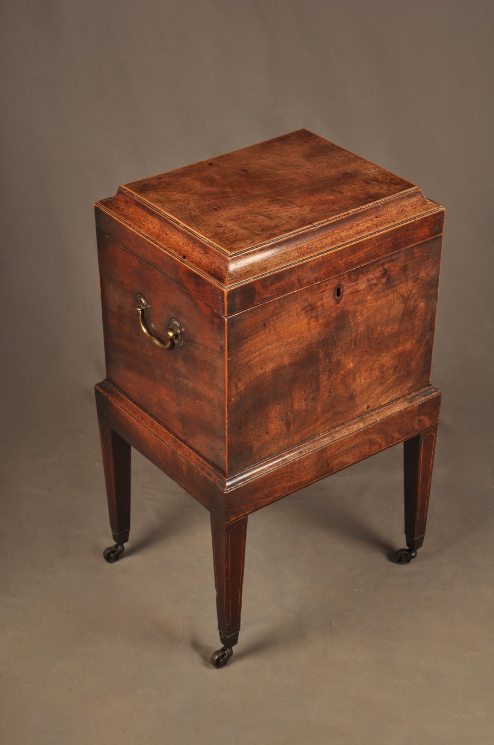 A Sheraton style mahogany cellarette featuring a hinged sarcophagus top, brass side handles, and square tapered legs on brass castors.