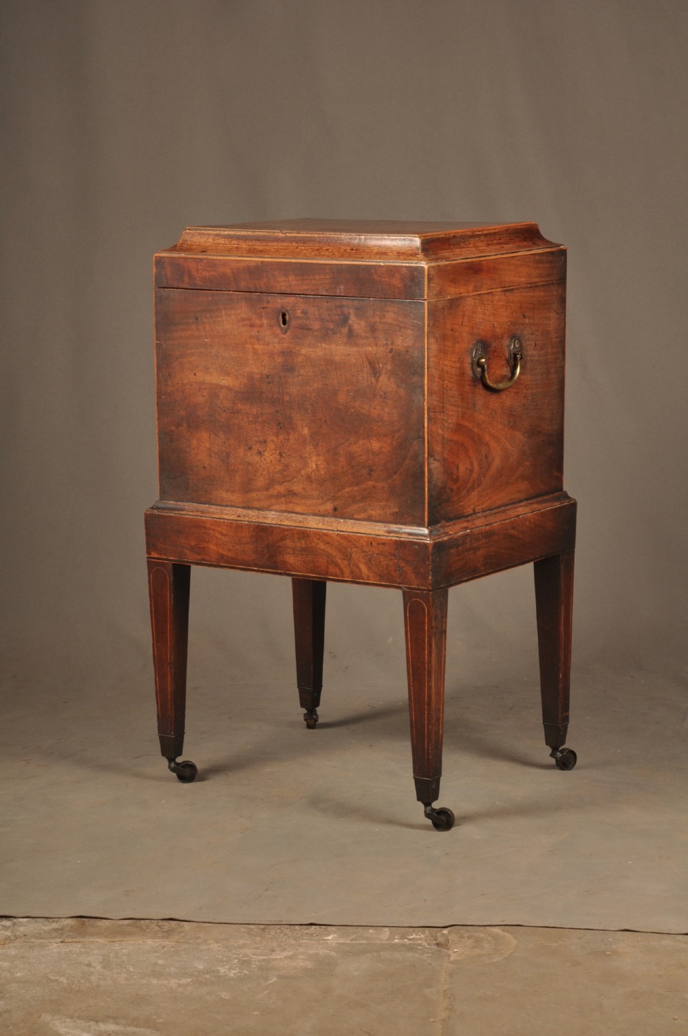 A Sheraton style mahogany cellarette featuring a hinged sarcophagus top, brass side handles, and square tapered legs on brass castors. View II